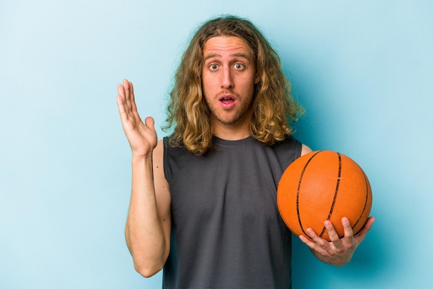 Junger kaukasischer Mann, der Basketball spielt, isoliert auf blauem Hintergrund, überrascht und schockiert