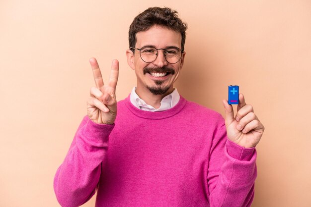 Junger kaukasischer Mann, der Batterien isoliert auf beigem Hintergrund hält und die Nummer zwei mit den Fingern zeigt.