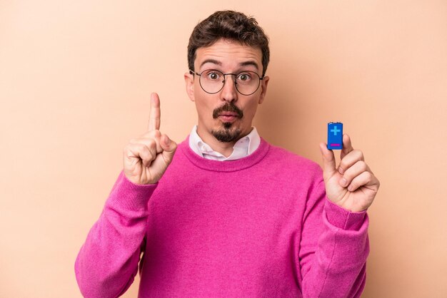 Junger kaukasischer Mann, der Batterien isoliert auf beigem Hintergrund hält und eine großartige Idee hat, Konzept der Kreativität.