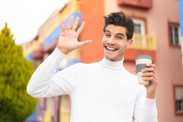 Junger kaukasischer mann, der draußen einen kaffee zum mitnehmen hält und mit der hand mit glücklichem ausdruck salutiert