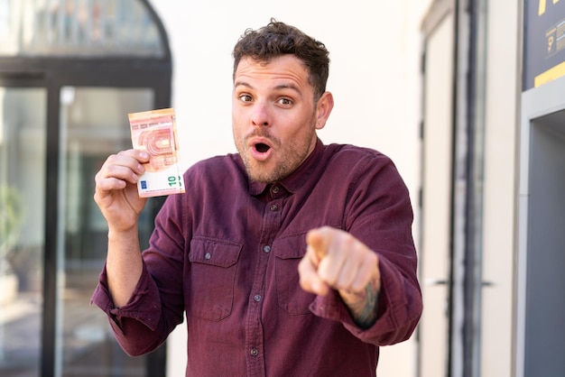 Foto junger kaukasischer mann, der draußen viel geld nimmt, überrascht und zeigt nach vorne