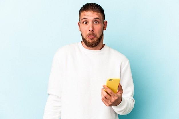 Junger kaukasischer Mann, der ein auf blauem Hintergrund isoliertes Handy hält, zuckt mit den Schultern und öffnet die Augen verwirrt.