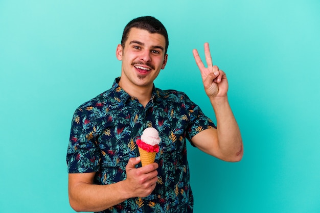 Junger kaukasischer Mann, der ein Eis isst, isoliert auf blauem Hintergrund, fröhlich und sorglos, das ein Friedenssymbol mit den Fingern zeigt.