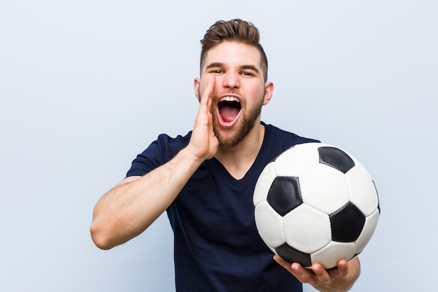 Junger kaukasischer Mann, der ein Fußballschreien aufgeregt zur Front hält.