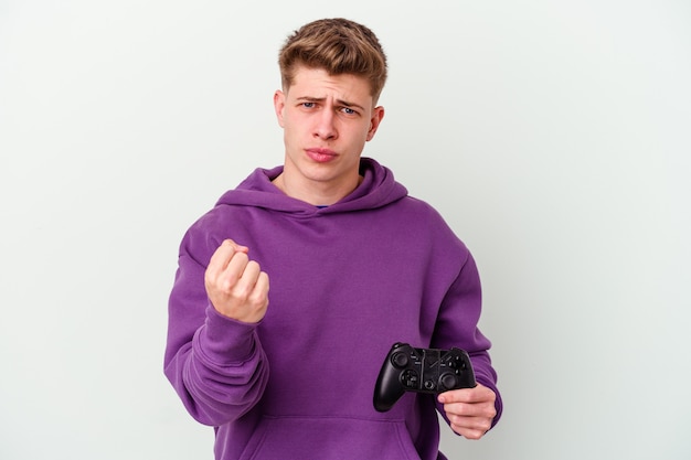 Junger kaukasischer Mann, der ein Gamepad lokalisiert auf weißer Wand hält, die Faust nach vorne zeigt, aggressiver Gesichtsausdruck