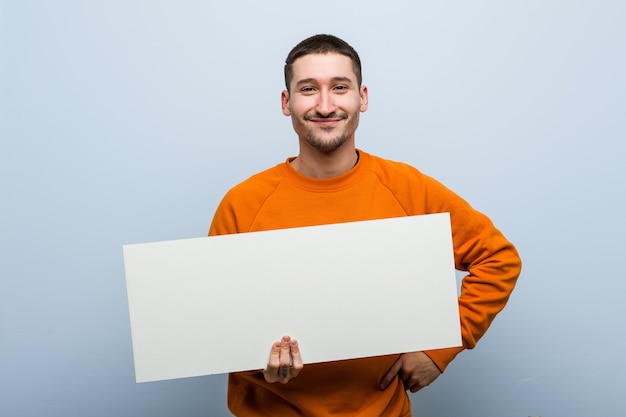 Junger kaukasischer Mann, der ein Plakat hält, das mit verschränkten Armen zuversichtlich lächelt.