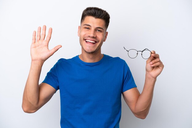 Junger kaukasischer Mann, der eine Brille isoliert auf weißem Hintergrund hält und mit der Hand mit glücklichem Ausdruck salutiert