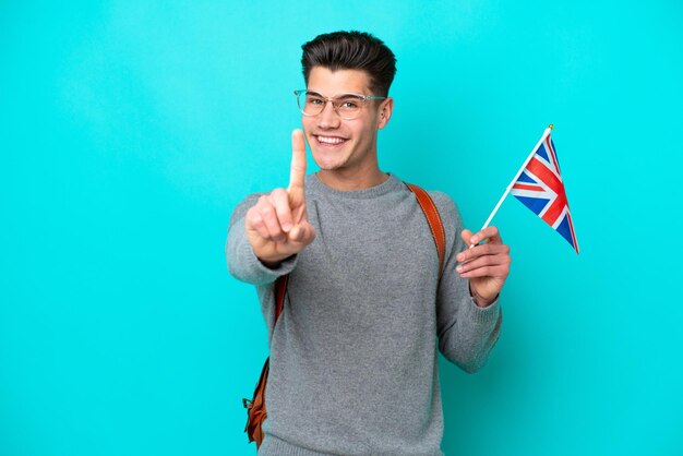 Junger kaukasischer Mann, der eine Flagge des Vereinigten Königreichs hält, die auf blauem Hintergrund isoliert ist und einen Finger zeigt und hebt