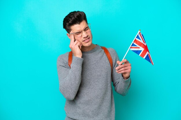 Junger kaukasischer Mann, der eine Flagge des Vereinigten Königreichs isoliert auf blauem Hintergrund mit Kopfschmerzen hält