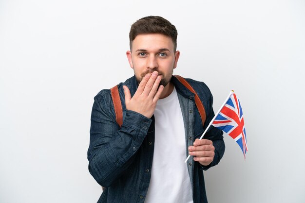 Junger kaukasischer Mann, der eine Flagge des Vereinigten Königreichs isoliert auf weißem Hintergrund hält, glücklich und lächelnd, den Mund mit der Hand bedeckend