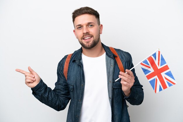 Junger kaukasischer Mann, der eine Flagge des Vereinigten Königreichs isoliert auf weißem Hintergrund hält und mit dem Finger zur Seite zeigt