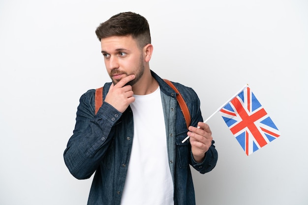 Junger kaukasischer Mann, der eine Flagge des Vereinigten Königreichs isoliert auf weißem Hintergrund hält und zur Seite schaut und lächelt