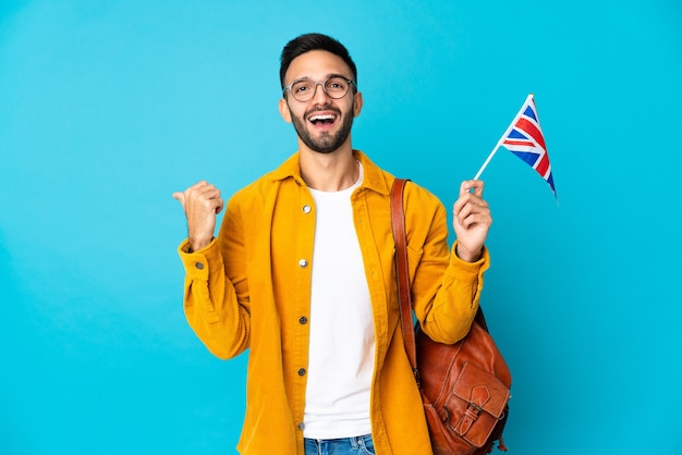 Junger kaukasischer Mann, der eine Flagge des Vereinigten Königreichs lokalisiert auf gelber Wand hält, die zur Seite zeigt, um ein Produkt zu präsentieren