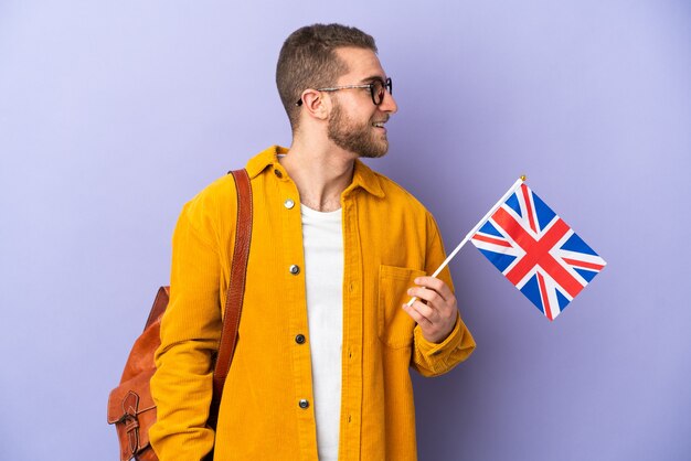 Junger kaukasischer Mann, der eine Flagge des Vereinigten Königreichs lokalisiert auf lila Wand schauender Seite hält