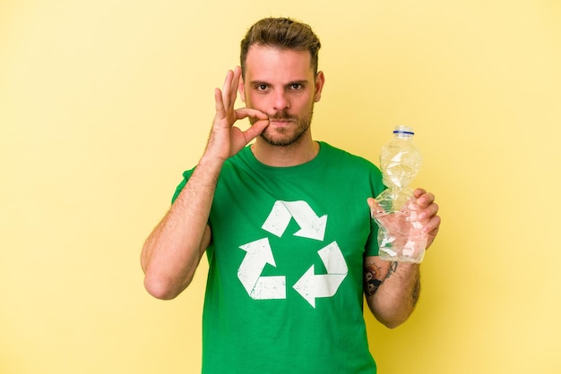 Junger kaukasischer mann, der eine flasche plastik hält, um isoliert auf gelbem hintergrund zu recyceln, mit den fingern auf den lippen, die ein geheimnis bewahren.