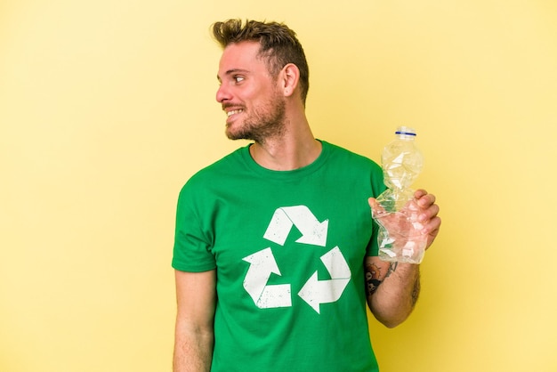 Junger kaukasischer Mann, der eine Flasche Plastik hält, um isoliert auf gelbem Hintergrund zu recyceln, sieht lächelnd, fröhlich und angenehm beiseite.