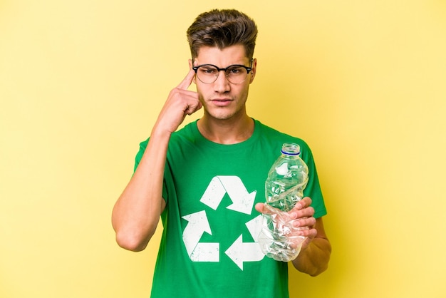 Junger kaukasischer mann, der eine flasche plastik zum recyceln hält, isoliert auf gelbem hintergrund, der mit dem finger auf die tempel zeigt, denkend, konzentriert auf eine aufgabe.