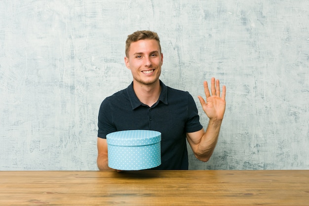 Junger kaukasischer Mann, der eine Geschenkbox auf einer Tabelle lächelt nette darstellende Nr. Fünf mit den Fingern hält.