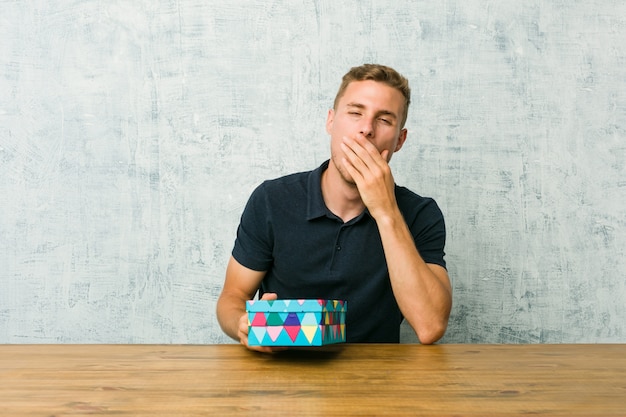 Junger kaukasischer Mann, der eine Geschenkbox auf einer Tabelle zeigt einen müden Gestenbedeckungsmund mit der Hand gähnend hält.