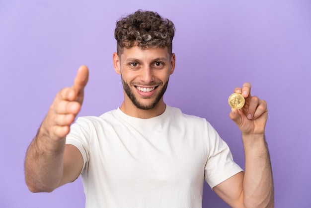 Junger kaukasischer Mann, der eine isolierte Bitcoin auf violettem Hintergrund hält und sich die Hände schüttelt, um ein gutes Geschäft abzuschließen