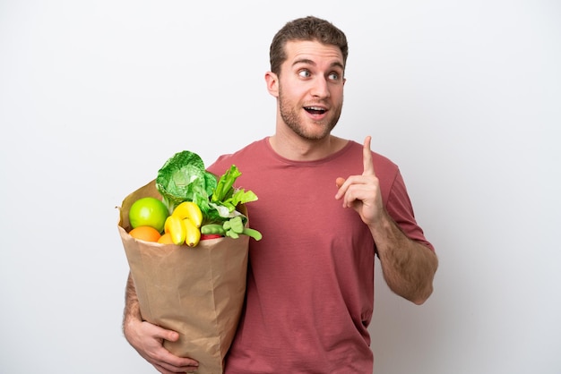 Junger kaukasischer Mann, der eine Lebensmitteleinkaufstasche lokalisiert auf weißem Hintergrund hält und eine Idee denkt, die den Finger nach oben zeigt