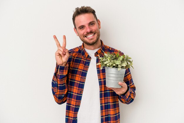 Junger kaukasischer Mann, der eine Pflanze isoliert auf weißem Hintergrund hält, fröhlich und sorglos, zeigt ein Friedenssymbol mit den Fingern