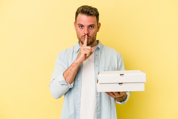 Junger kaukasischer mann, der eine pizza isoliert auf gelbem hintergrund hält, ein geheimnis bewahrt oder um stille bittet.