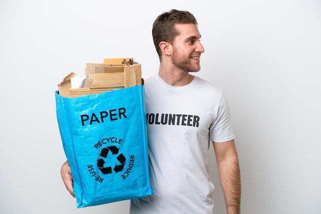 Junger kaukasischer Mann, der eine Recycling-Tasche voll Papier hält, um lokalisiert auf weißem Hintergrund, der Seite schaut, zu recyceln