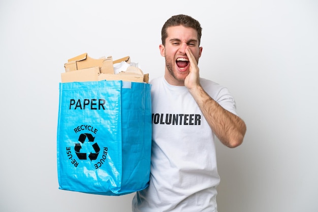 Junger kaukasischer Mann, der eine Recyclingtüte voller Papier hält, um sie isoliert auf weißem Hintergrund zu recyceln, schreit mit weit geöffnetem Mund