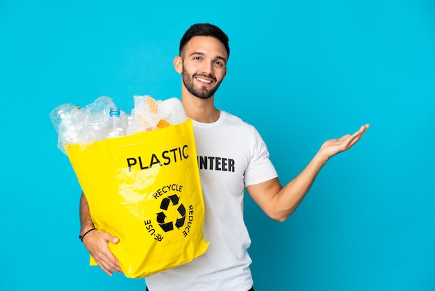 Junger kaukasischer Mann, der eine Tasche voller Plastikflaschen hält, um auf blauem Hintergrund isoliert Hände zu recyceln, die Hände zur Seite für das Einladen kommen, zu kommen