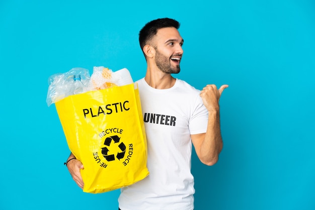 Junger kaukasischer Mann, der eine Tasche voller Plastikflaschen hält, um auf blauem Hintergrund isoliert zu recyceln, in der Absicht, die Lösung zu realisieren, während ein Finger anhebt