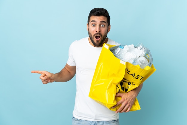 Junger kaukasischer Mann, der eine Tasche voller Plastikflaschen hält, um isoliert auf blauer Wand überrascht und zeigende Seite zu recyceln