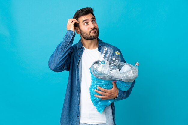 Junger kaukasischer Mann, der eine Tasche voller Plastikflaschen hält, um isoliert auf blauer Wand zu recyceln, die Zweifel und mit verwirrendem Gesichtsausdruck hat