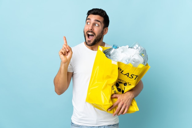 Junger kaukasischer Mann, der eine Tasche voller Plastikflaschen hält, um isoliert auf der blauen Wand zu recyceln, die beabsichtigt, die Lösung zu realisieren, während ein Finger anhebt