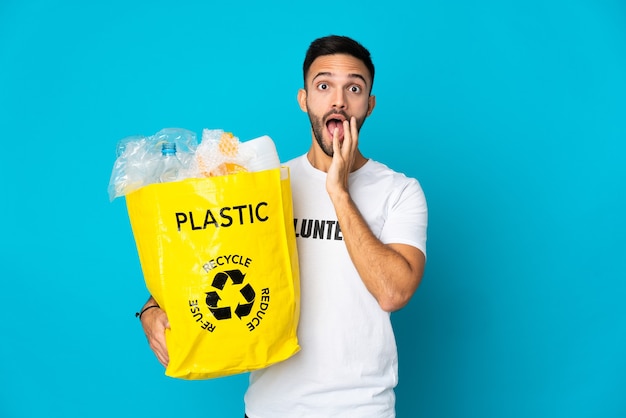 Junger kaukasischer Mann, der eine Tasche voller Plastikflaschen hält, um lokalisiert auf blauem Hintergrund mit Überraschung und schockiertem Gesichtsausdruck zu recyceln