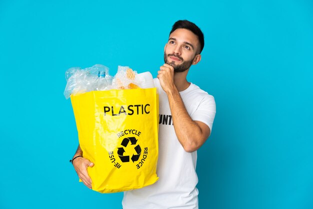 Junger kaukasischer Mann, der eine Tasche voller Plastikflaschen hält, um lokalisiert auf blauem Hintergrund und nach oben zu recyceln