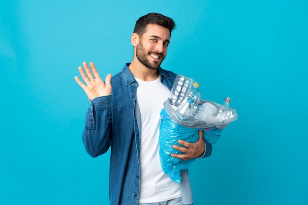 Junger kaukasischer Mann, der eine Tasche voller Plastikflaschen hält, um lokalisiert auf der blauen Wand, die mit Hand mit glücklichem Ausdruck salutiert, zu recyceln