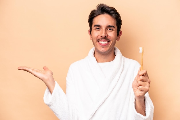 Junger kaukasischer Mann, der einen Bademantel trägt und eine Zahnbürste hält, isoliert auf beigem Hintergrund, zeigt einen Kopierraum auf einer Handfläche und hält eine andere Hand an der Taille.