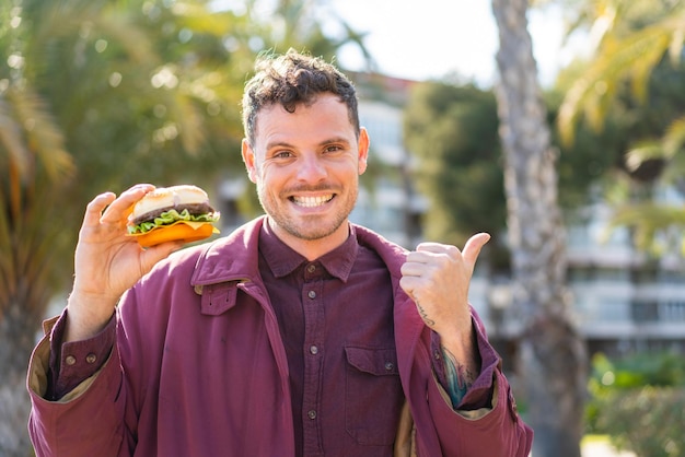 Junger kaukasischer mann, der einen burger im freien hält und zur seite zeigt, um ein produkt zu präsentieren