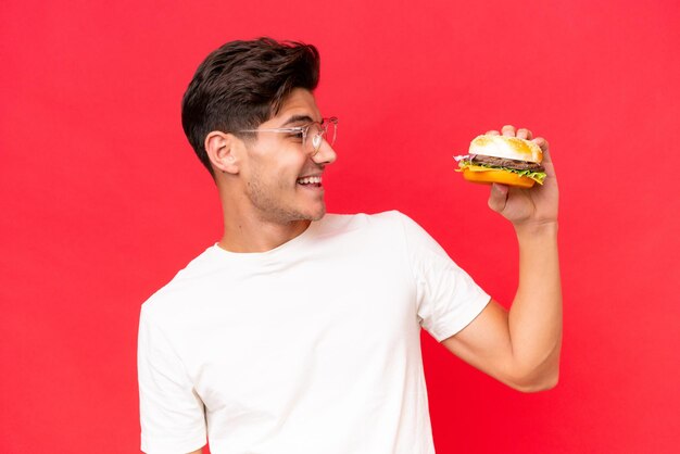 Junger kaukasischer Mann, der einen Burger lokalisiert auf rotem Hintergrund mit glücklichem Ausdruck hält