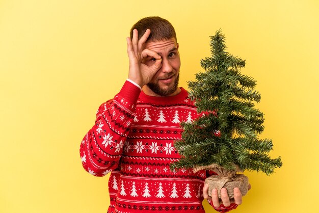 Junger kaukasischer Mann, der einen kleinen Baum zu Weihnachten kauft, isoliert auf gelbem Hintergrund, aufgeregt, die Geste auf dem Auge zu halten.