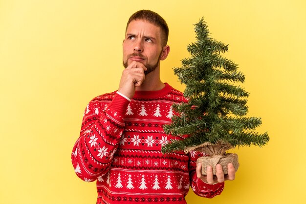 Junger kaukasischer Mann, der einen kleinen Baum zu Weihnachten kauft, isoliert auf gelbem Hintergrund, der seitlich mit zweifelhaftem und skeptischem Ausdruck schaut.