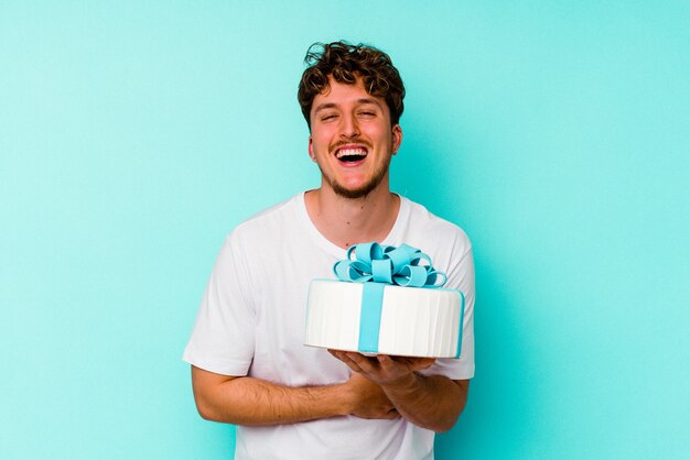 Junger kaukasischer Mann, der einen Kuchen lokalisiert auf der blauen Wand lacht und Spaß hat hält.