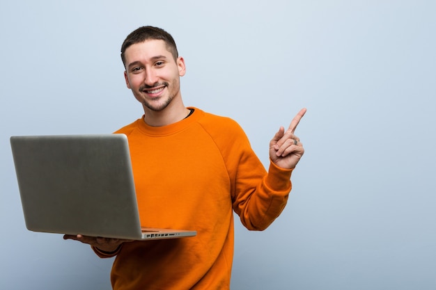 Junger kaukasischer Mann, der einen Laptop hält, der fröhlich mit Zeigefinger weg zeigt.