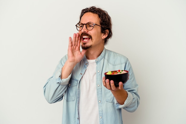 Junger kaukasischer Mann, der einen Ramen isst, der auf weißer Wand lokalisiert wird, die Handfläche nahe geöffnetem Mund schreit und hält.