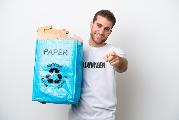 Junger kaukasischer Mann, der einen Recycling-Beutel voller Papier hält, um isoliert auf weißem Hintergrund zu recyceln, der mit glücklichem Ausdruck nach vorne zeigt