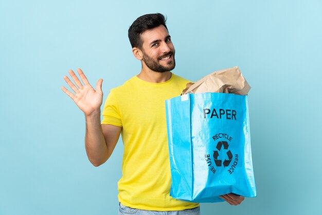 Junger kaukasischer Mann, der einen Recyclingbeutel lokalisiert auf blauer Wand hält, die mit Hand mit glücklichem Ausdruck salutiert