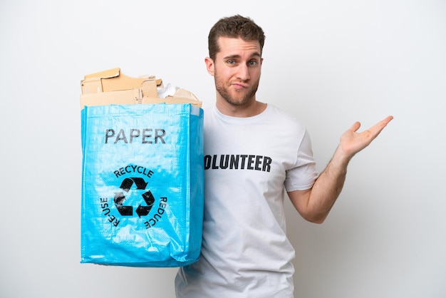 Junger kaukasischer Mann, der einen Recyclingbeutel voll Papier hält, um lokalisiert auf weißem Hintergrund mit Zweifeln beim Aufheben der Hände zu recyceln