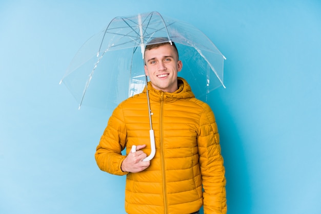 Junger kaukasischer Mann, der einen Regenschirm glücklich, lächelnd und fröhlich hält.