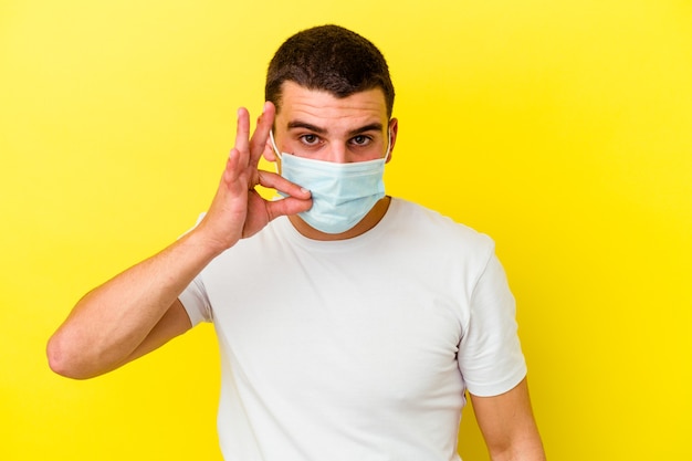 Junger kaukasischer Mann, der einen Schutz für Coronavirus trägt, lokalisiert auf gelbem Hintergrund mit den Fingern auf den Lippen, die ein Geheimnis halten.